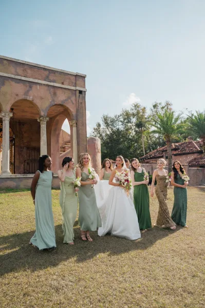 Bridesmaid group photo of the bride with bridesmaids in mismatched green dresses holding bouquets under string lights by stucco arches