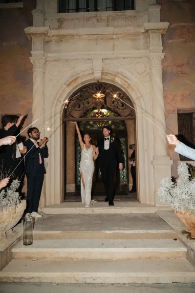 Wedding exit with sparkler wedding send off as bride in beaded slit gown raises her arm beside groom in tux under stone arch at night