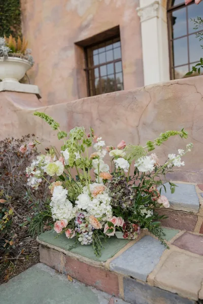 Wedding floral arrangement of blush roses, carnations, and hydrangea with greenery on stone steps by a stucco wall and windows