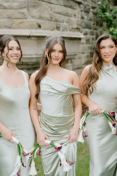 Bridesmaid portrait of three women in sage green bridesmaid dresses holding calla lily bouquets against a stone wall with greenery