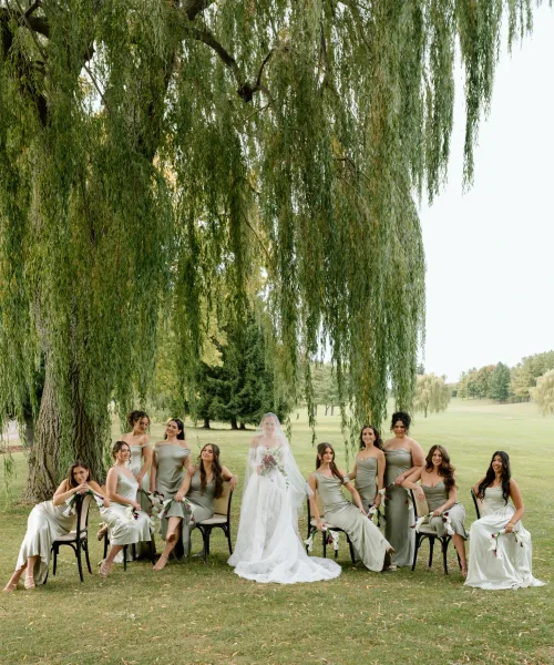 Bride with bridesmaids seated in a bridal party portrait, holding burgundy and white bouquets beneath a weeping willow on a park lawn
