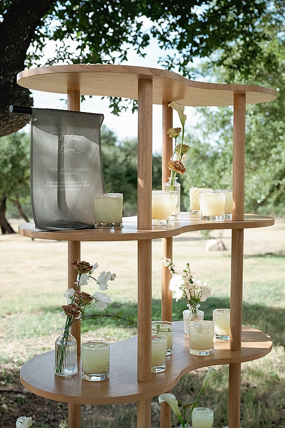 Wedding welcome drinks on a wood display shelf with cocktail glasses and printed bar menu sign, styled with white flowers, on a sunny lawn