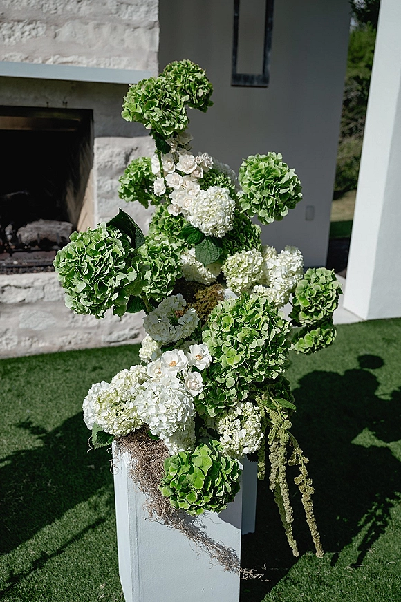 Wedding floral arrangement of green and white hydrangeas with white roses and hanging amaranthus on a pedestal by a stone fireplace outdoors