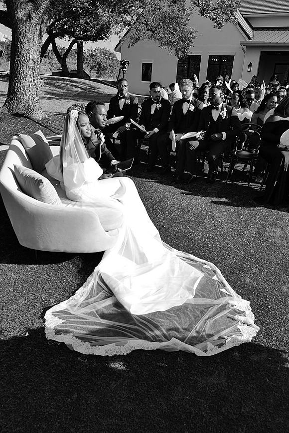 Wedding ceremony moment with bride and groom seated on a sofa, her long lace-trim veil spread on the lawn as guests watch under a large tree