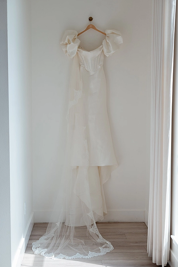 Wedding dress on hanger with corset bodice and puff sleeves, paired with a lace veil, hanging by window light against a white wall