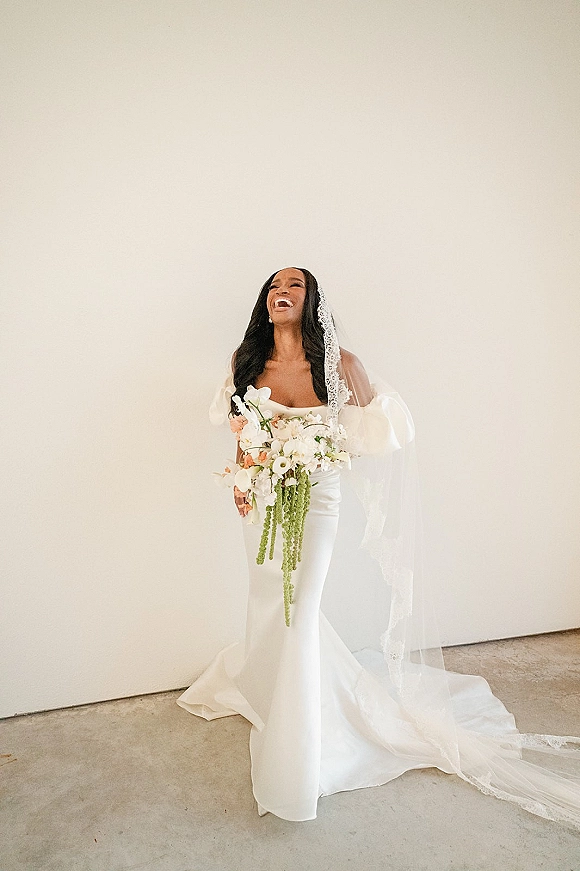 Bridal portrait of a laughing bride in a strapless wedding dress holding a cascading greenery bouquet, lace veil and pearl earrings by a white wall