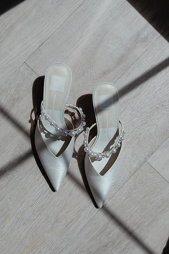 Bridal shoes with pearl wedding shoes ankle straps in white satin, pointed toes on a light wood floor with soft window shadows