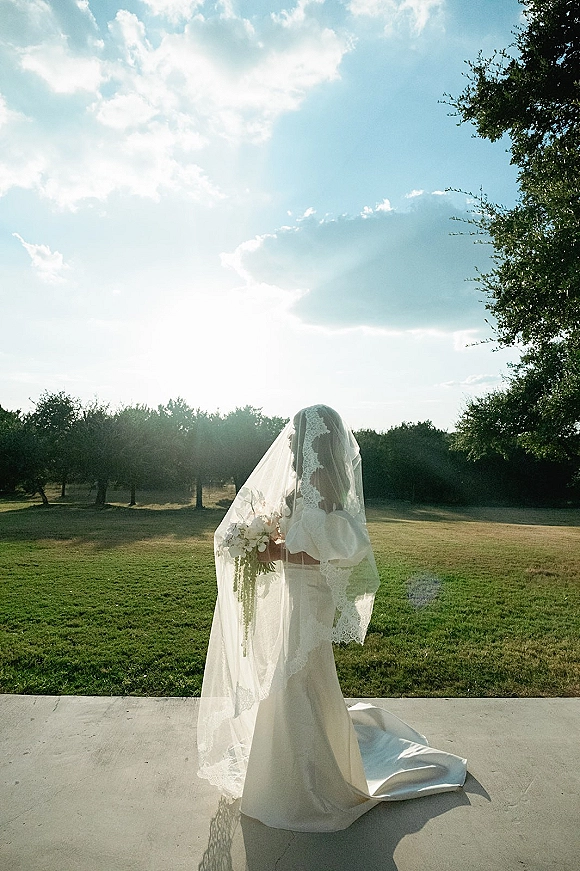 Bridal portrait of a bride holding bouquet under a long lace-edged veil, off-the-shoulder gown and train in a sunlit field with trees