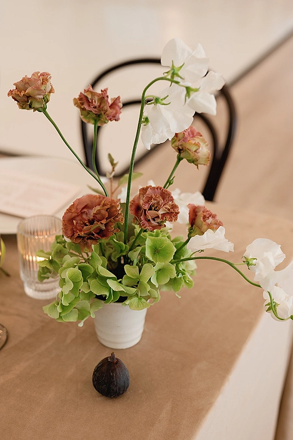 Wedding centerpiece with sweet peas and green hydrangea in a white ceramic vase beside a fig on a brown paper runner at the reception table
