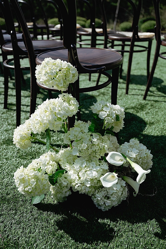 Ceremony aisle decor with wedding aisle flowers in white hydrangeas and calla lilies beside black wooden chairs on a green garden lawn