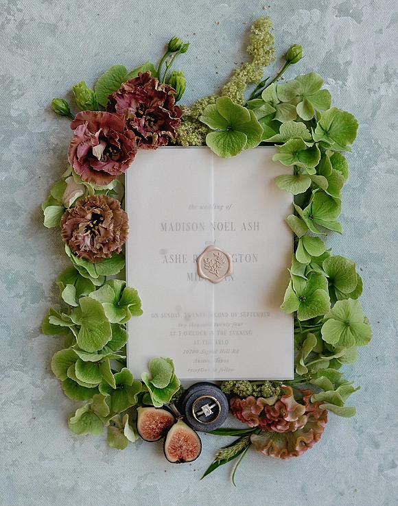 Wedding invitation suite with vellum wedding invitation, wax seal, hydrangea blooms, figs, and engagement ring on a light gray stone surface