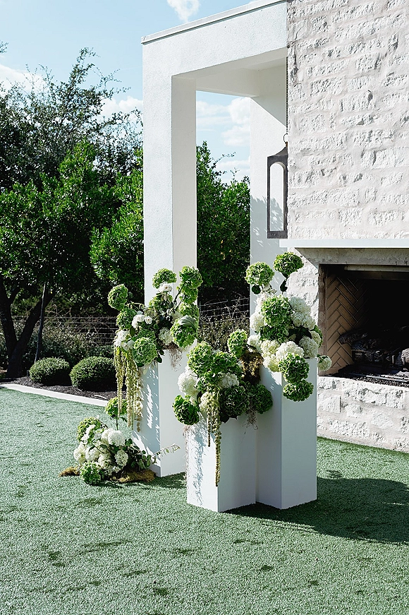 Wedding ceremony flowers on white pedestal plinths with green-and-white arrangements and hanging greenery against a white stucco fireplace outdoors