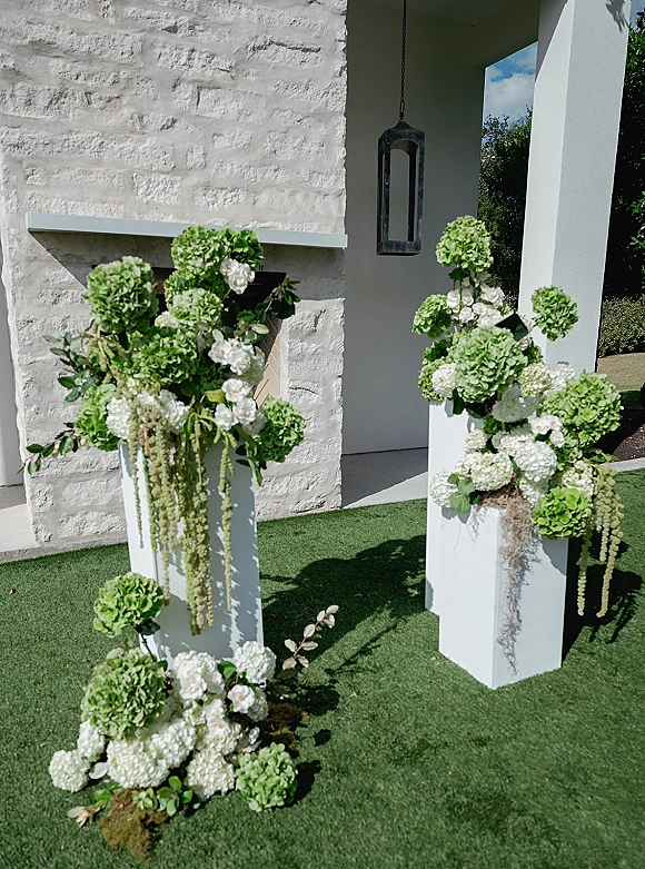 Ceremony floral pedestals with wedding pedestal arrangements of hydrangeas, white roses, and greenery on white plinths by stone columns and lantern