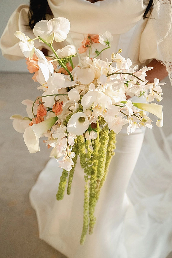 Bridal bouquet with orchid wedding bouquet blooms, calla lilies and hanging amaranthus, held by a bride in a lace sleeve indoors