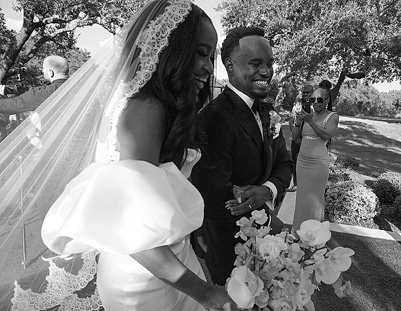 Wedding recessional as bride and groom walking down a garden walkway, smiling with bouquet and lace-trim veil as guests cheer behind