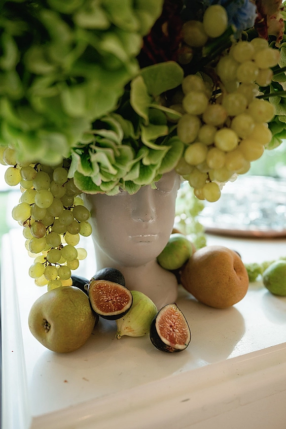 Fruit centerpiece with grape centerpiece styling, green hydrangea, figs, pears and citrus surrounding a white mannequin head vase on a windowlit table