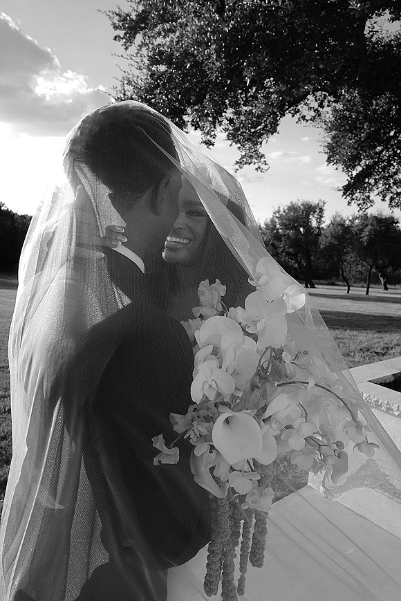 Wedding couple portrait with bride and groom under veil, bride holding a cascading orchid bouquet on an outdoor lawn by trees and sky