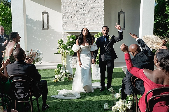 Wedding recessional as bride and groom walking aisle hand in hand past cheering guests, her off-the-shoulder gown and lantern-lined florals outside fireplace backdrop