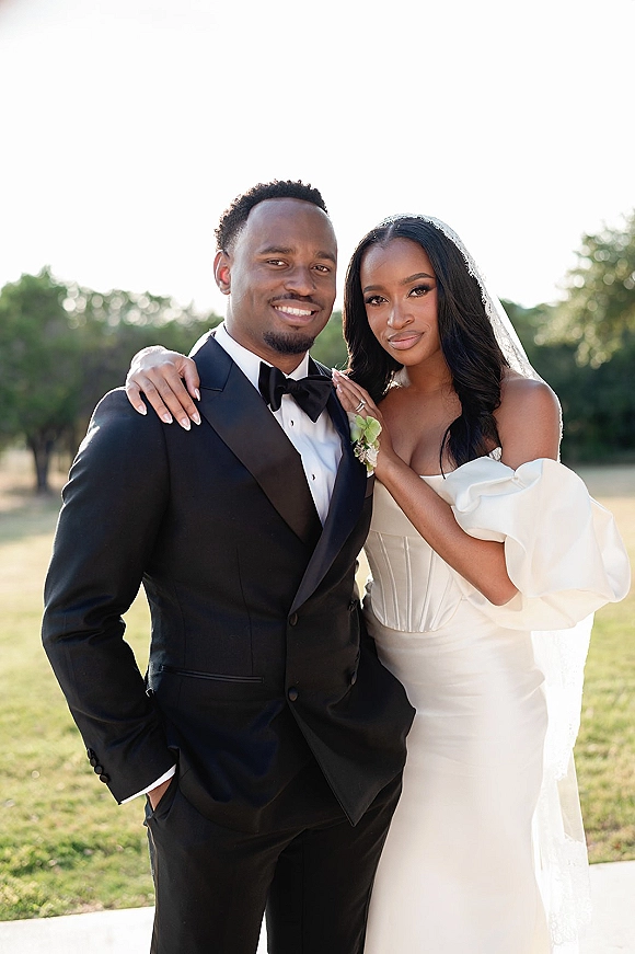 Couple portrait of bride and groom outdoors, bride’s hand on groom showing her wedding ring, wearing veil and black tuxedo on a lawn