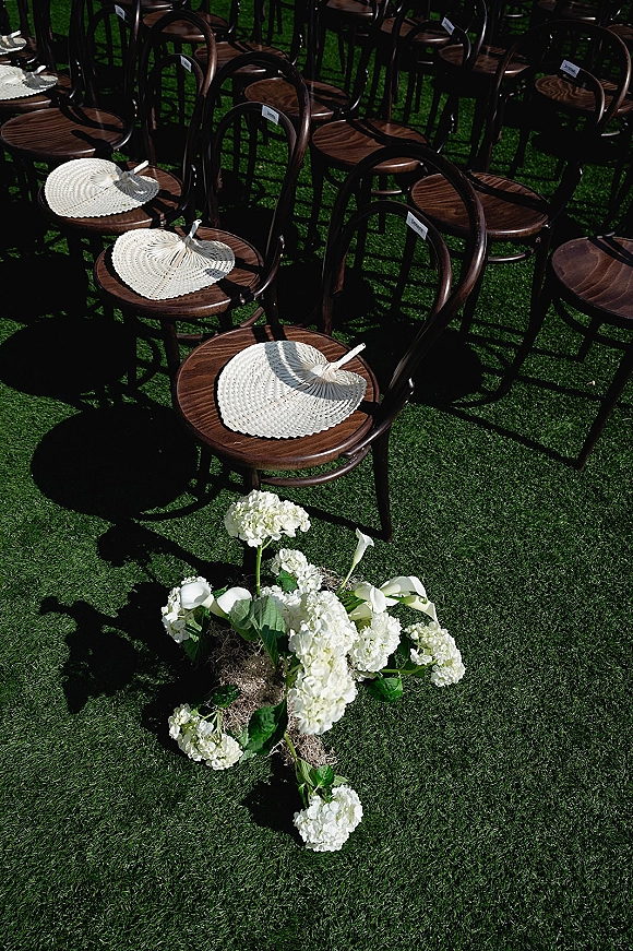 Ceremony seating with outdoor wedding chairs in dark wood, each with a woven hand fan, beside white hydrangea and calla lilies on a sunlit lawn