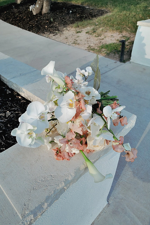 Wedding bouquet with white orchids and calla lilies, blush blooms and greenery, resting on a stone ledge beside a concrete walkway