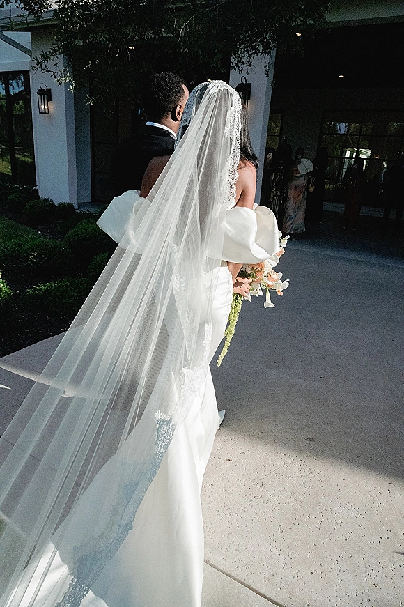 Wedding kiss portrait of bride and groom kiss, her long lace veil flowing behind as they walk past white doors with guests nearby