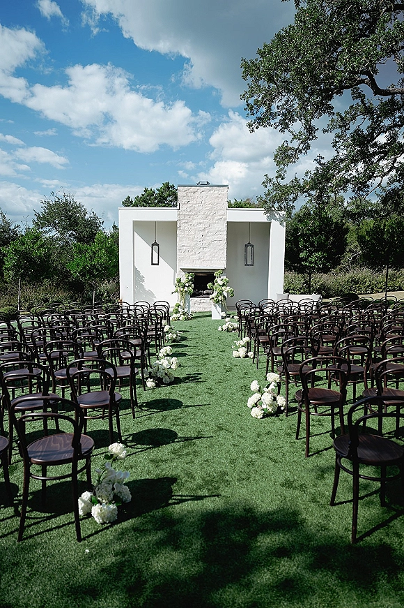 Ceremony setup for an outdoor wedding ceremony with white aisle flowers, dark wood chairs, and lanterns framing a stone fireplace altar