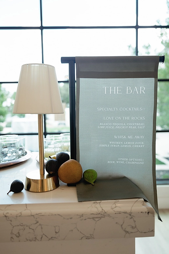 Bar menu sign wedding bar menu displayed on a marble bar top beside a tabletop lamp and cocktail glasses by a large window with greenery