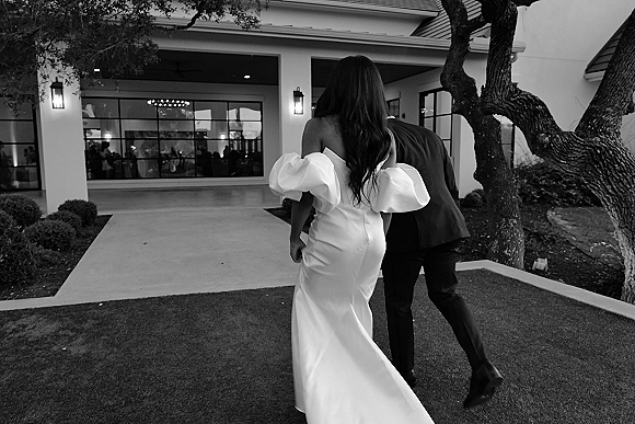 Newlywed couple, bride and groom walking hand in hand toward the venue entrance, bride’s train trailing beside large windows and lanterns