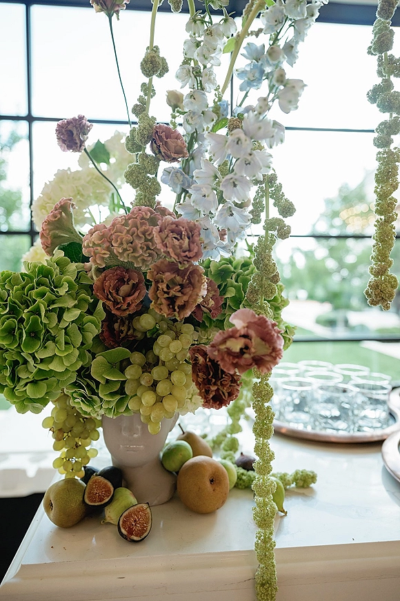 Wedding centerpiece with hydrangeas and hanging amaranthus, roses, and fruit on a pedestal vase, set by a window wall with greenery