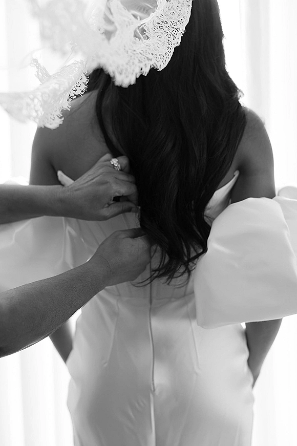 Bride getting ready as a bridesmaid zips the wedding dress zipper back, off-the-shoulder sleeves and lace veil glowing in window light