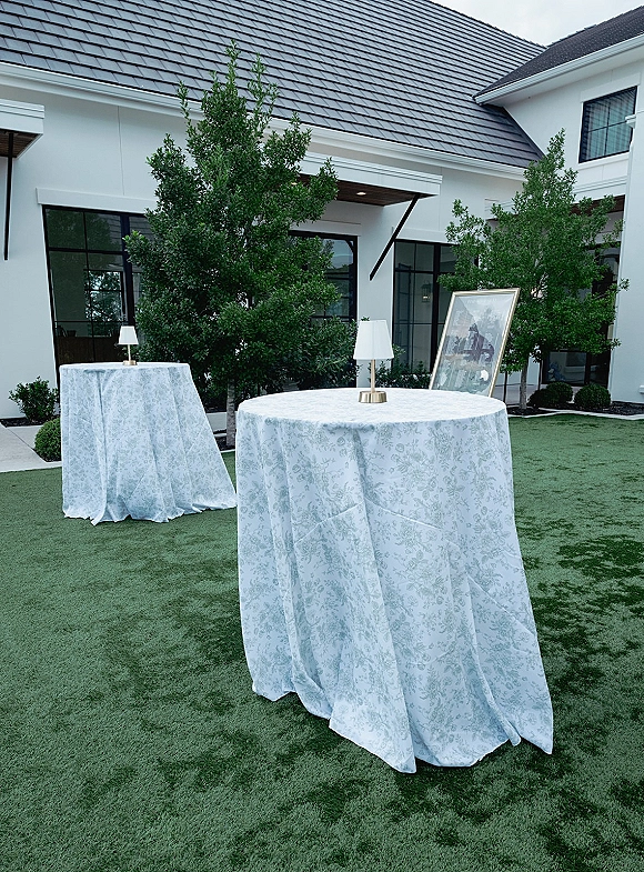 Cocktail table decor with outdoor cocktail hour setup featuring patterned linens and a table lamp, arranged on a lawn by a modern white building