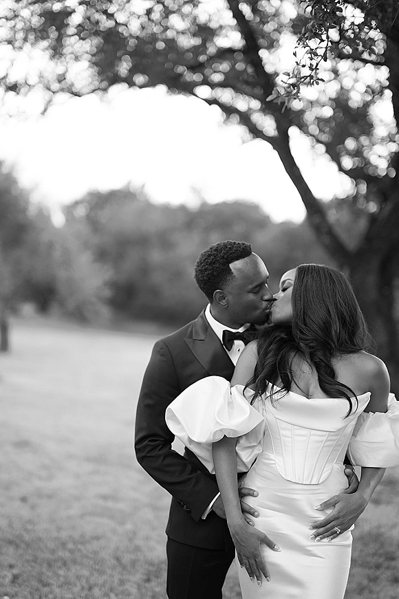 Wedding kiss portrait of bride and groom kissing as he embraces her, in a grassy park with trees; she wears a puff sleeve gown
