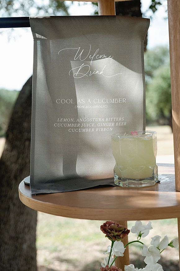 Wedding bar menu fabric sign with calligraphy lettering beside a cocktail glass on a wooden bar shelf in sunlit outdoor greenery
