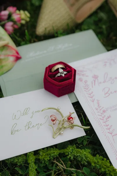 Wedding rings with engagement ring detail on invitation cards beside a velvet ring box, pink flowers and greenery on mossy grass backdrop