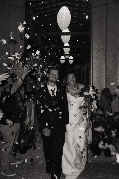 Wedding exit as bride in a strapless dress holds her bouquet beside groom in suit while guests toss petals under string lights in a dark hallway