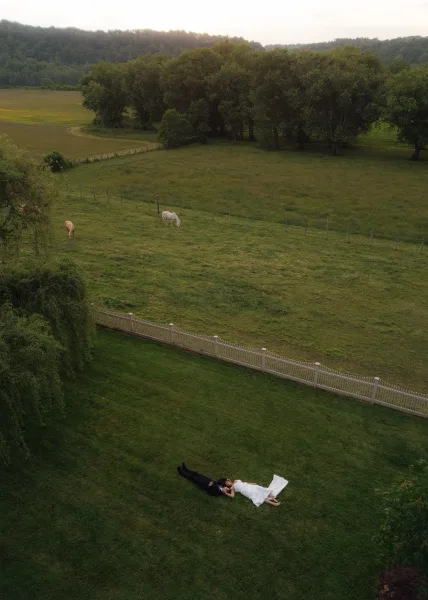 Couple portrait of bride and groom lying down on green lawn, her wedding dress train spread out, with white fence and horses behind