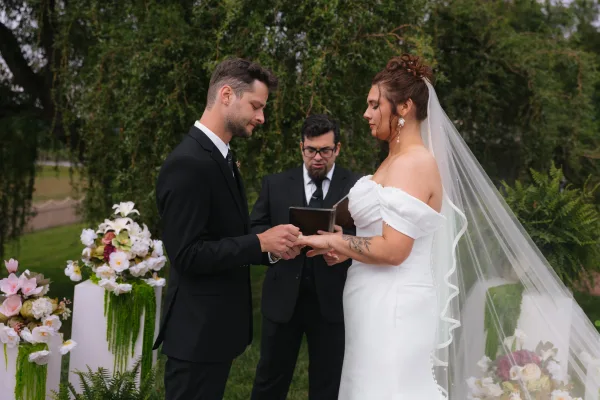 Wedding ring exchange as groom in black tuxedo places ring on bride in off-shoulder gown and veil during outdoor garden ceremony