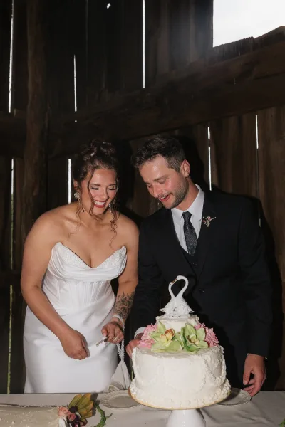 Wedding cake cutting as bride in strapless dress and groom in black suit slice a two-tier floral cake with swan topper before a rustic barn wall