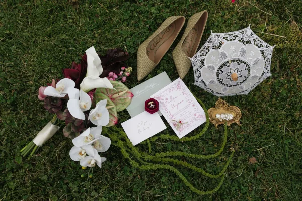 Invitation suite wedding invitation flat lay on a grass lawn with sage envelopes, RSVP card, velvet ring box, bouquet, heels, and lace parasol