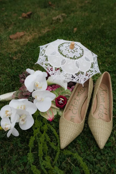 Wedding details flatlay with champagne wedding heels, engagement ring in a velvet box, lace parasol, and orchids on grass lawn