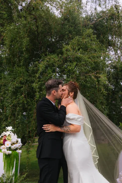 Wedding kiss portrait of bride and groom kissing, her veil flowing as she cups his face, bouquet on floral pedestal in garden greenery
