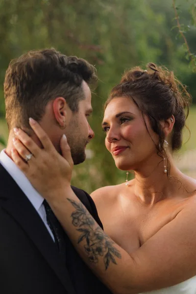 Couple portrait of bride touching groom’s face in a wedding couple close up, showing her ring and earrings against blurred greenery backdrop