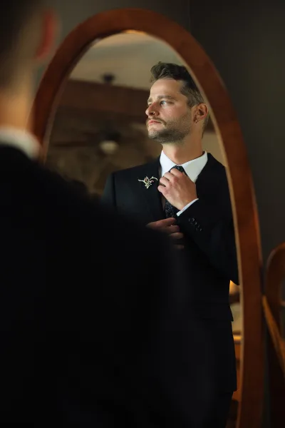 Groom portrait of a bearded groom adjusting his dark patterned tie in a mirror, wearing a black suit and boutonniere in warm indoor light