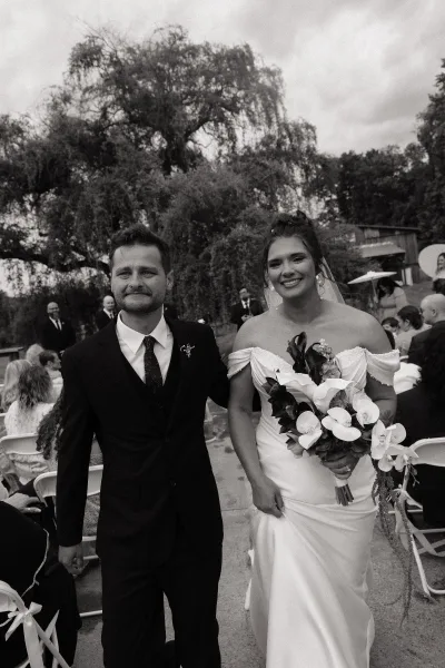 Wedding couple portrait of bride and groom walking aisle, bride holding an orchid bouquet with veil, guests and trees under cloudy sky