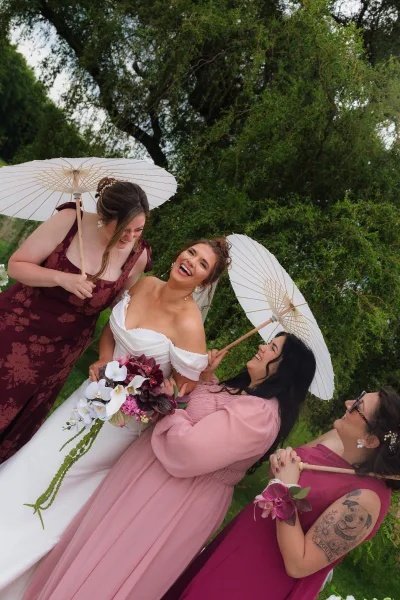 Bride with bridesmaids laughing together, holding paper parasols and orchid bouquet, in burgundy dresses on a green garden lawn under overcast sky