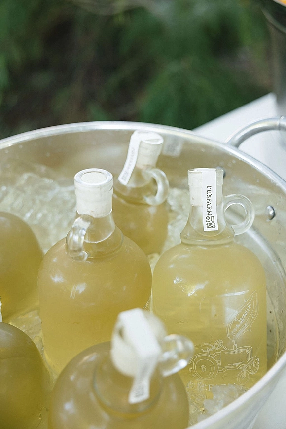 Wedding drink station with glass growlers on ice, labeled bottle tags and metal ice bucket on a white tablecloth outdoors