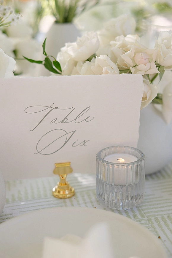 Wedding table number with calligraphy table numbers in a gold holder beside a glass votive candle, white roses and greenery on tablecloth