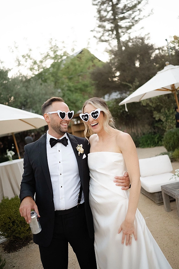 Couple portrait of bride and groom laughing in heart-shaped sunglasses, hugging on an outdoor patio lounge under white umbrellas