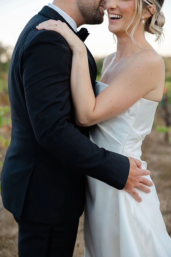 Wedding couple portrait of groom kissing the bride’s cheek as she laughs, showing her engagement ring in a field under open sky